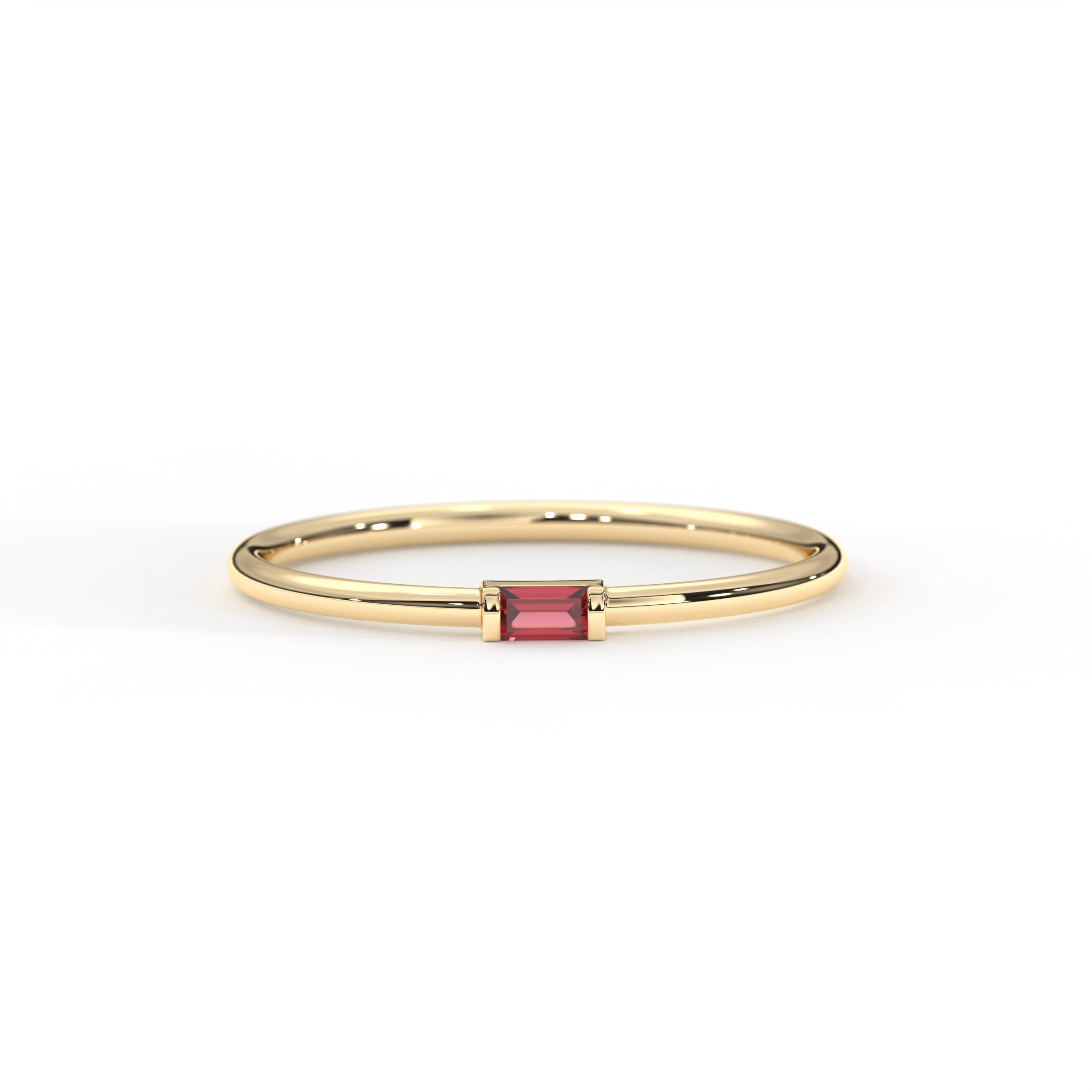 A delicate solid gold band ring with a small, rectangular, baguette red garnet gemstone set in the center.