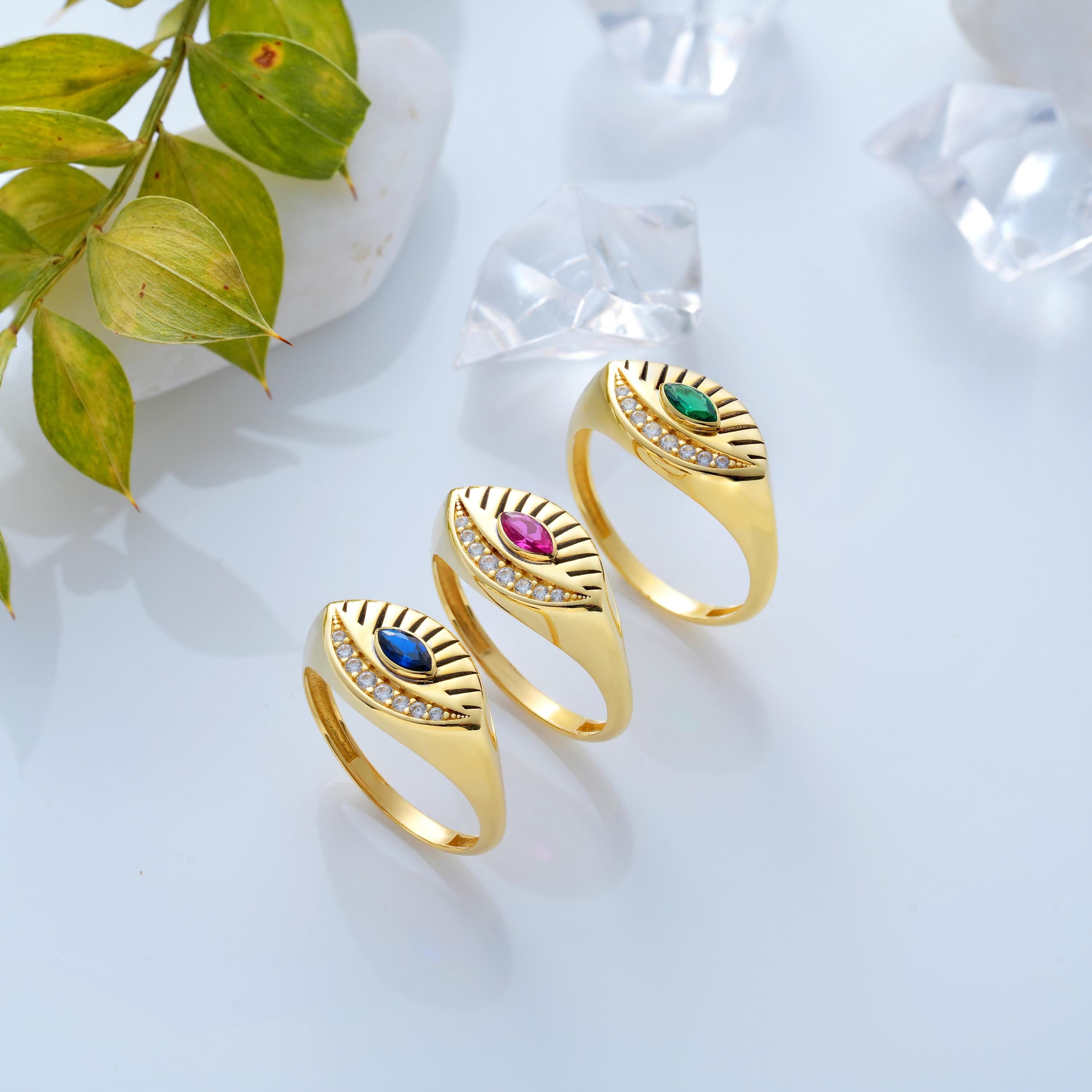 Three solid gold evil eye rings on white surface with marquise emerald, sapphire, and ruby colored stones with engraved rays and diamond pave accents